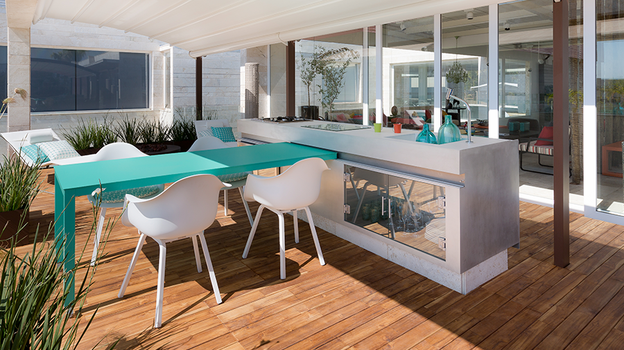 Imagem de uma área gourmet com bancada branca, mesa azul e cadeiras brancas com folhas ao redor. Piso de deck de madeira e ao fundo, portas de correr brancas