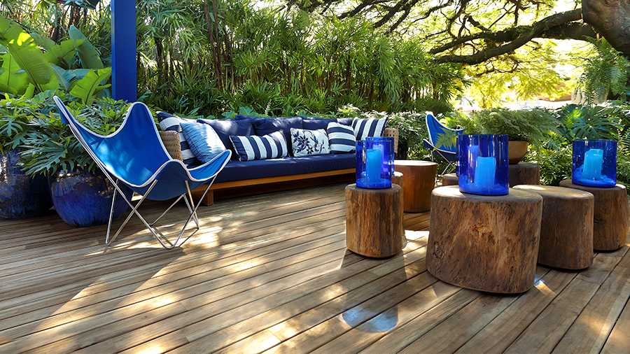 Ambiente externo de descanso com sofás, e cadeiras em azul, deck de madeira e árvores ao fundo.