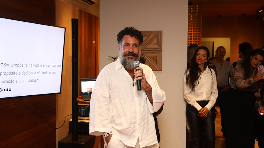 Designer Pedro Franco, com camisa branca e microfone na mão, fala para diversas pessoas, ao lado de uma televisão com frase de Buda e amostras de revestimento de madeira ao fundo.
