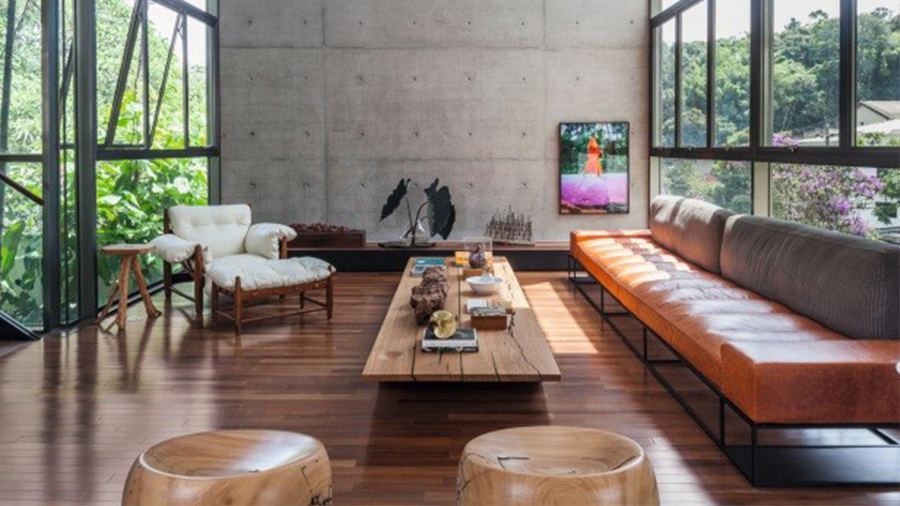 Imagem de uma sala com uma parede de concreto adornada com um quadro e piso de madeira. Dentro da sala, há um sofá caramelo, uma poltrona branca, uma mesa e dois bancos de madeira. As duas paredes laterais são feitas de vidro.