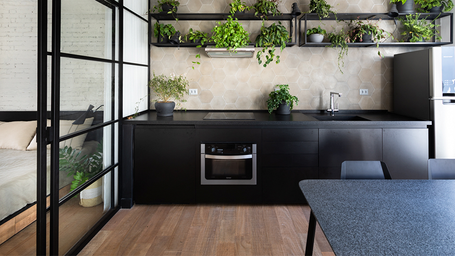 Cozinha estilo industrial, com parede de cimento queimado, piso de madeira, estruturas em ferro e móveis preto. Plantas verdes complementam a decoração do ambiente