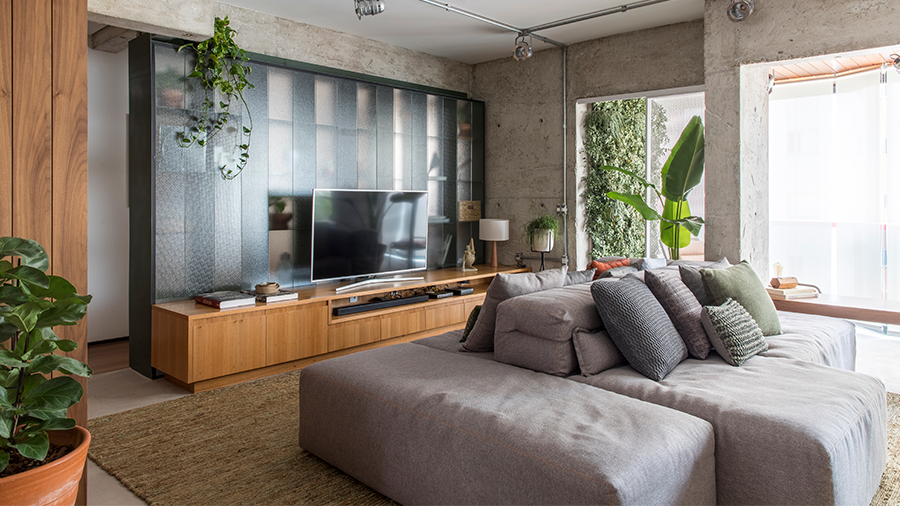 Imagem de uma sala com paredes pintadas no estilo cimento queimado e decorações em madeira, incluindo um hack que suporta a TV e vários objetos. Na frente, há um sofá cinza, e o espaço é cercado por plantas.