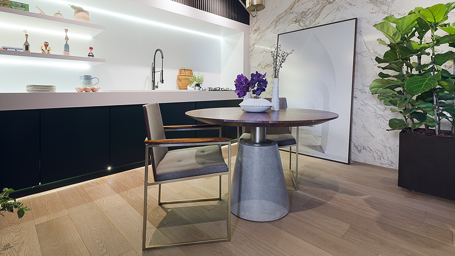 Ambiente com piso de madeira engenheirada Carvalho da Masterpiso, mesa redonda no centro, parede revestida com textura mármore, vaso grande de planta e móvel branco e preto. 