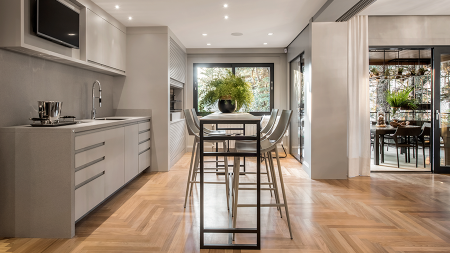 Cozinha com piso de madeira, móveis e decoração  em tons claros de creme e branco e porta de vidro ao fundo sendo possível visualizar o jardim.