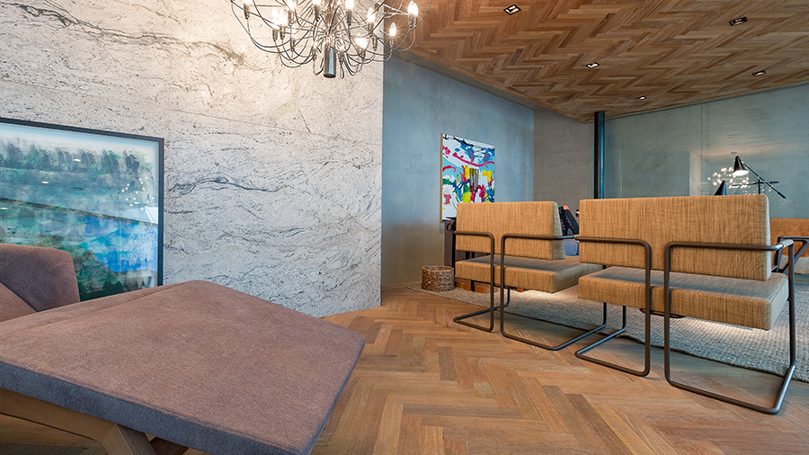 Ambiente com piso e revestimento de teto de madeira chevron, móveis e decoração em tons próximos de madeira, lustre e quadro abstrato na parede