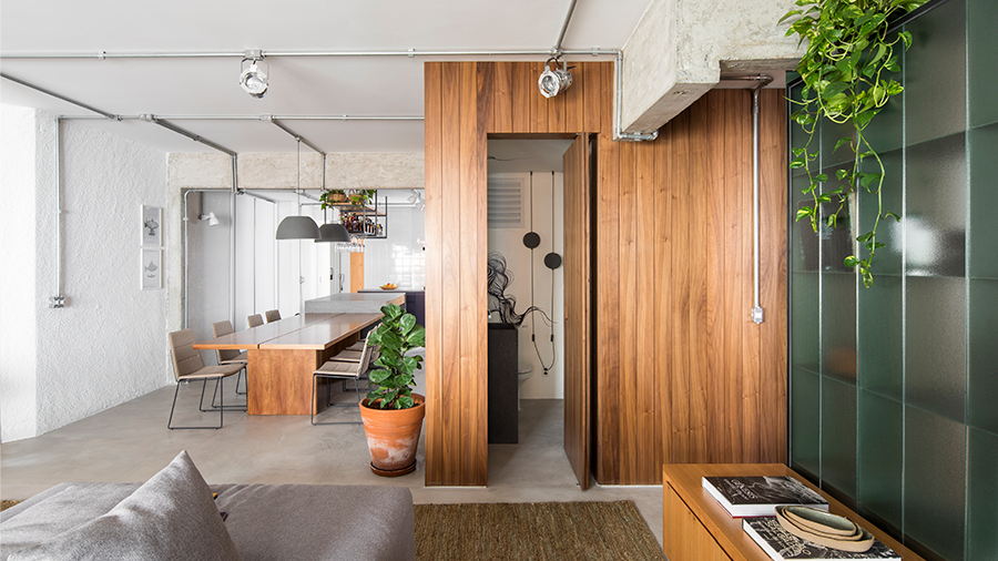 Imagem de uma casa com conceito aberto, contendo sofá, tapete, armário em madeira e uma estante. Na parede de fundo, destaca-se uma parede também em madeira, com um vaso sobre uma mesa rodeada de cadeiras.