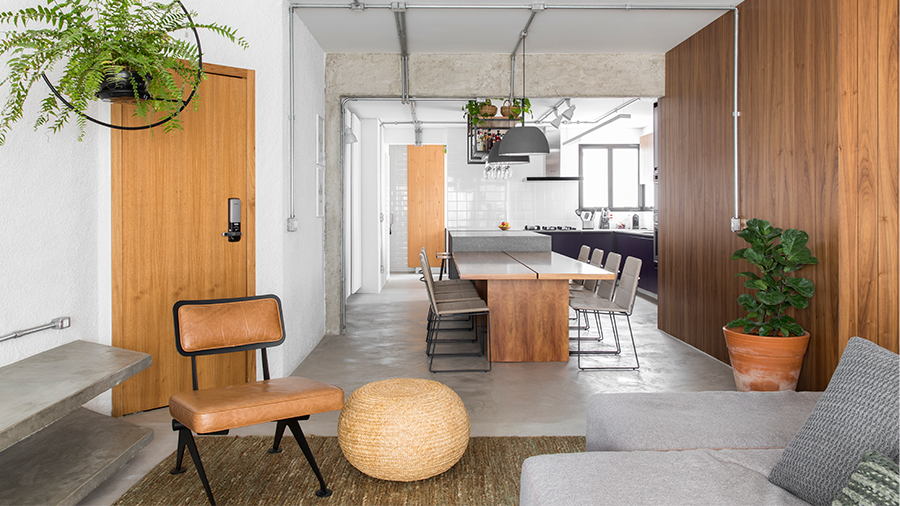 Imagem de um ambiente em tons de cinza e marrom. O espaço apresenta um sofá com uma cadeira ao lado, e ao fundo, uma parede de madeira com uma mesa e cadeiras na frente, integrando-se a uma cozinha.