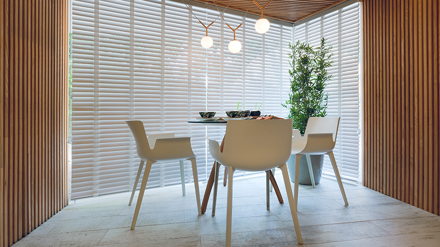  Imagem de uma sala minimalista com paredes e teto de madeira Brise Carvalho, um lustre triplo, uma mesa redonda com quatro cadeiras e um vaso grande com folhagens ao lado.