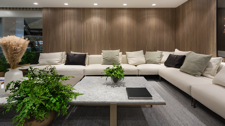 Imagem de uma ampla sala com um sofá em L branco, com almofadas de diversas cores, um tapete e uma mesa de centro com um vaso de planta em cima e ao lado. Ao fundo, uma parede de madeira.