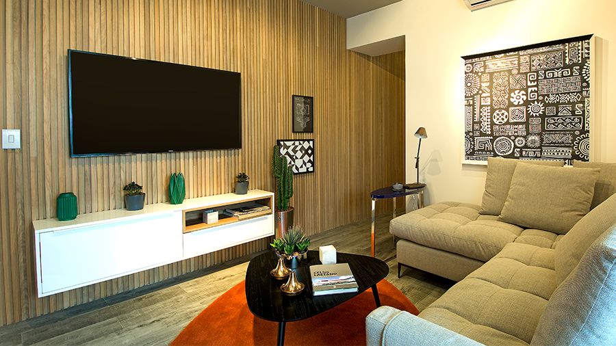 Imagem de uma sala com um sofá em L, um tapete com uma mesa de centro com objetos. Na frente, um armário e uma TV suspensa na parede. Uma das paredes é de madeira, a outra é branca, com um ar-condicionado e um quadro. 