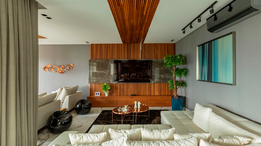 Sala com sofá branco, mesa de centro sobre tapete, armário e parede de madeira com TV e um vaso de plantas ao lado. O teto possui uma faixa em madeira e as paredes exibem um quadro e uma escultura.