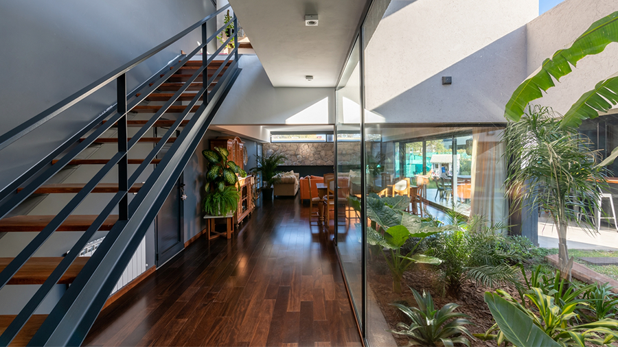 Imagem do corredor de uma casa com piso de madeira Elegance Sucupira, com uma escada. Ao fundo, móveis e plantas complementam o ambiente. Ao lado, uma parede envidraçada com um jardim externo.