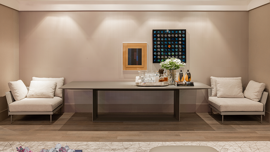  Imagem de um ambiente com cores claras, duas poltronas e uma mesa retangular entre elas com objetos concentrados em uma das pontas, dois quadros na parede e pido de madeira Sucupira.