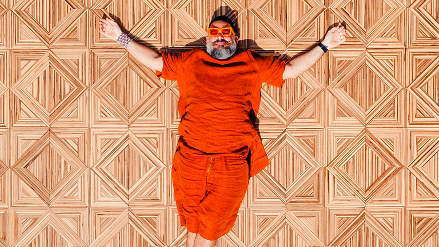 Imagem de um homem com camisa, calça e óculos laranja deitado com os braços abertos sobre um piso de madeira com paginação versailles.