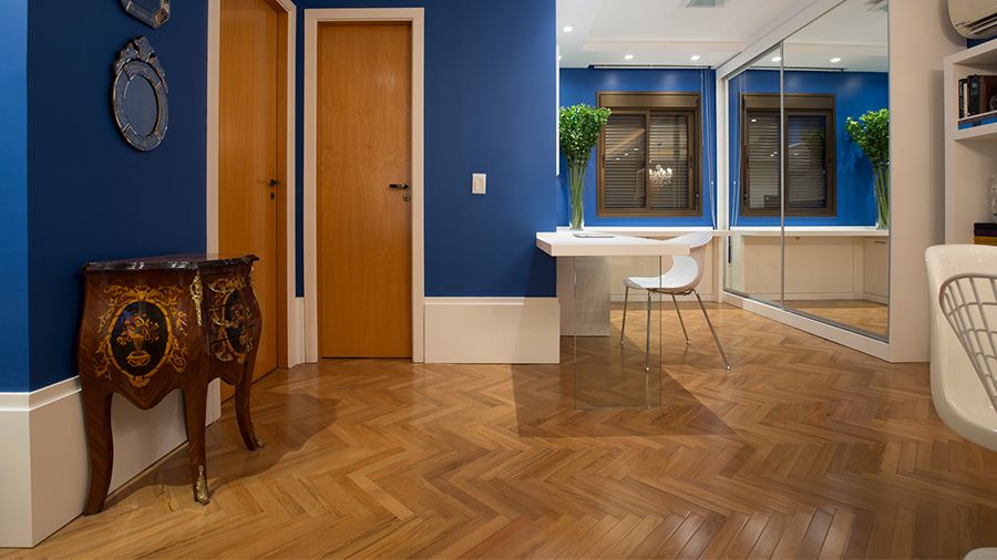 Imagem da parte interna de uma casa com piso de madeira Peroba Dourada envernizado. As paredes são azuis escuro, há duas portas, um aparador e ao fundo um armário com espelho e uma mesa com uma cadeira.
