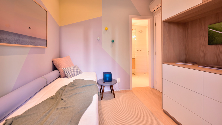  Imagem de um quarto com uma cama de solteiro encostada na parede, com uma manta e almofadas. À frente, há um armário de madeira, um banco ao lado e as paredes pintadas em tons pastéis com várias formas assimétricas.