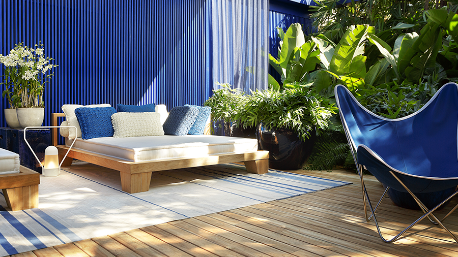 Imagem de um deck de madeira com uma espreguiçadeira, almofadas brancas e azuis sobre um tapete. Ao redor, uma poltrona azul e vasos com plantas, com a parede de fundo em azul e diversas plantas ao lado.