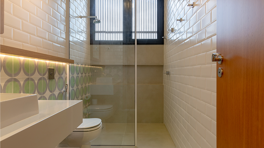 Imagem de um banheiro com pia e vaso sanitário de um lado. Metade da parede possui lajotas brancas e verdes, enquanto a outra metade é revestida com tijolos brancos. Do outro lado, a porta, ao fundo um box de vidro e uma janela.