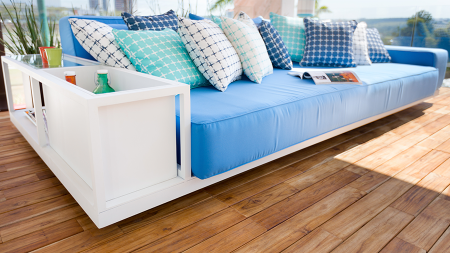magem de um sofá azul com almofadas coloridas, com um baú acoplado em um dos braços contendo objetos. O ambiente possui piso de madeira.