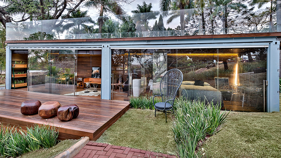 Imagem da área externa de uma casa com gramado, plantas e um deck de madeira Ipê Champagne. Ao fundo, destaca-se a casa com portas de vidro.