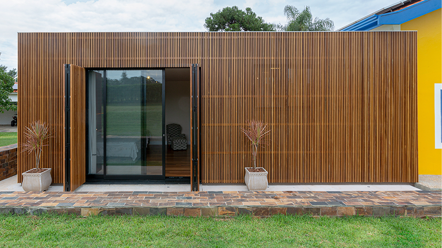 Imagem externa de uma casa com as paredes revestidas de madeira brise Ipê, apresentando portas de vidro. Ao lado, uma casa amarela, e à frente, há um gramado.
