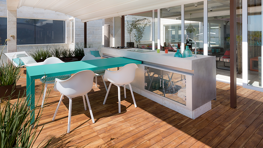 Imagem de um deck de madeira em uma área externa com um balcão branco decorado com objetos, uma mesa azul rodeada por cadeiras e plantas espalhadas pelo ambiente. Ao fundo, é possível ver janelas e portas de vidro que dão acesso a uma casa.