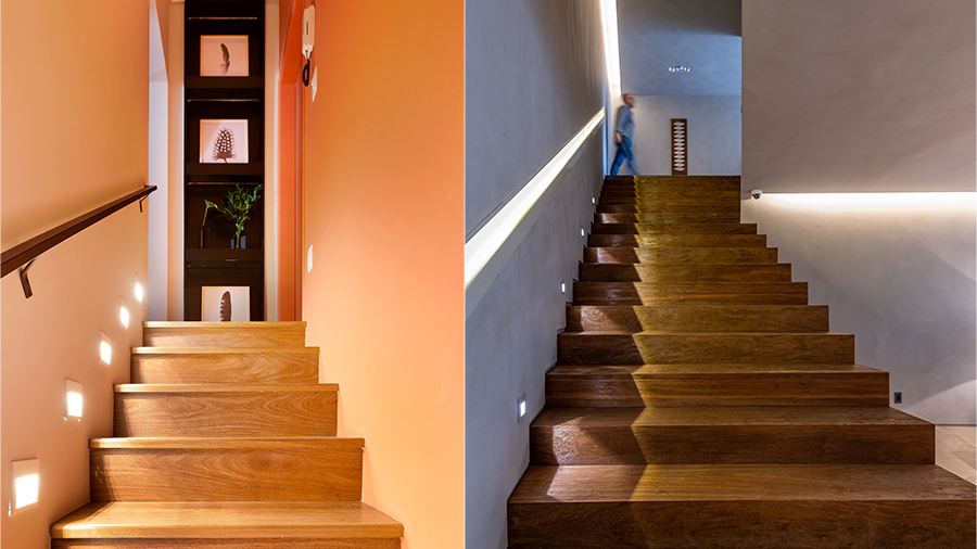Imagem de uma escada de madeira com corrimão, luminárias no chão e, no fim do corredor, quadros pendurados. Ao lado, uma escada de madeira com corrimão iluminado, luminárias de chão, e uma pessoa ao fundo.