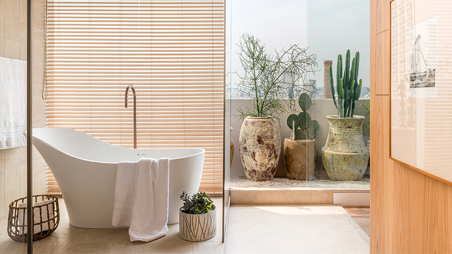 Imagem de uma banheira com uma torneira e uma toalha, acompanhada por um vaso e um cesto do lado de fora. Uma das paredes é de vidro, enquanto a outra apresenta um quadro. Ao fundo, há um jardim pequeno de inverno.