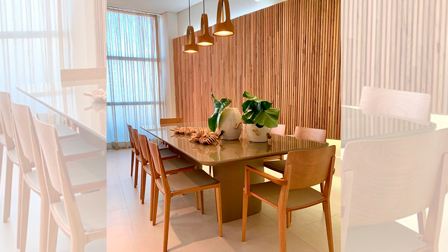 Sala de jantar com mesa de 8 lugares, com cadeiras de madeira, três lustres pendentes acima da mesa e parede com revestimento Brise de madeira freijó