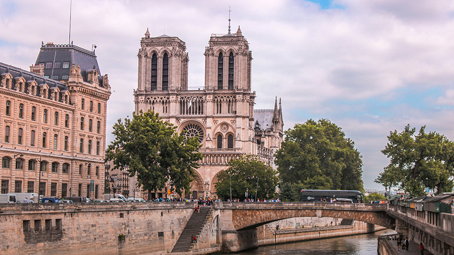  Imagem da Catedral de Notre-Dame exibindo sua arquitetura gótica vista de longe, com ruas e árvores ao redor, além de uma ponte à sua frente.