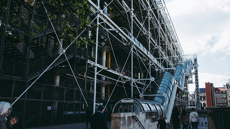 Imagem do Centro Georges Pompidou que exibe um túnel de acesso lateral, estruturas em formato de 'X' e uma arquitetura moderna. Ao redor, há diversas pessoas circulando.