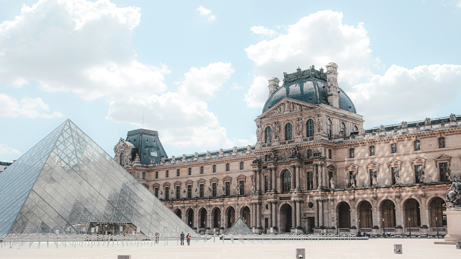 Imagem do Museu do Louvre exibindo sua arquitetura renascentista, com a pirâmide de vidro à sua frente e pessoas circulando ao redor.