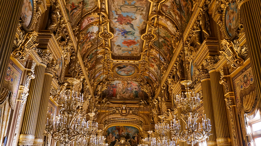  Imagem do interior da Ópera Garnier com o teto pintado, com diversos lustres enfileirados, colunas decoradas e estrutura luxuosa.