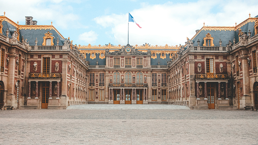 Imagem do Palácio de Versalhes exibindo sua arquitetura barroca, com tons em marrom e azul e uma bandeira pendurada ao centro.