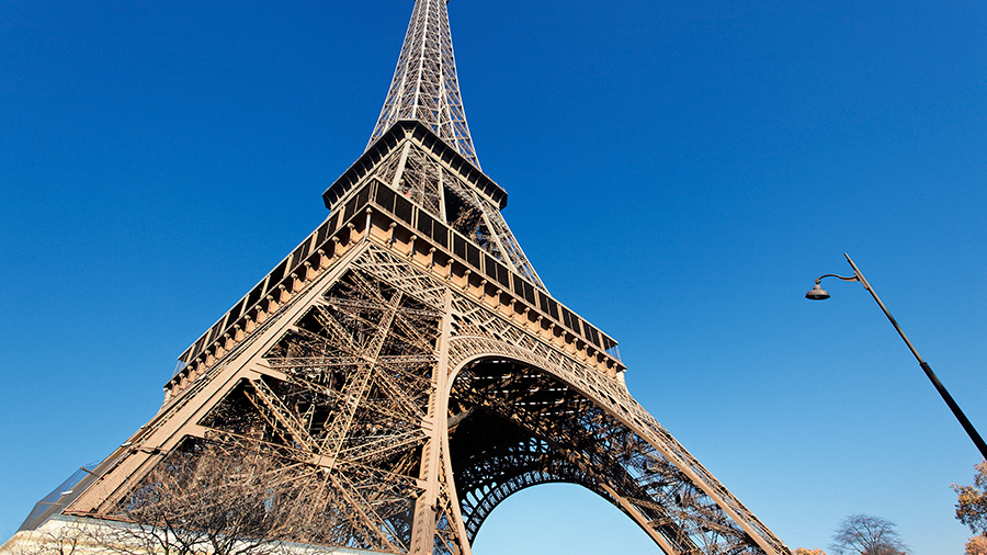 Imagem da Torre Eiffel capturada de baixo para cima, com várias árvores ao redor e o céu azul sem nuvens.