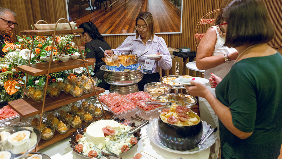 Imagem de uma mesa com bolos, frios, uma estrutura em forma de pirâmide com doces espalhados e uma variedade de comidas, com pessoas ao redor da mesa durante o encontro de revendas de 2024.
