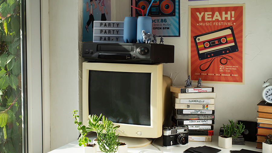 Imagem de uma mesa com a tela de um computador antigo, sobre ele um DVD e um pequeno letreiro, e ao redor, livros e plantas pequenas espalhadas. Na parede, há quadros.