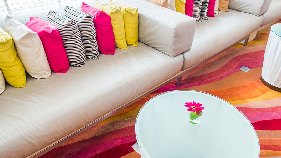 Imagem de um sofá claro comprido com almofadas coloridas. À frente, uma mesa circular e um tapete nas cores rosa, laranja, vermelho e bege.