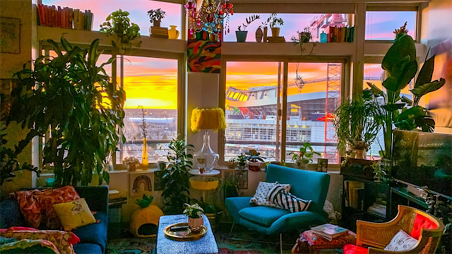 Imagem de uma sala com sofás com almofadas coloridas, poltronas também coloridas com almofadas, mesa de centro com um vaso, diversas plantas e objetos ao redor, e janelas ao fundo com vasos pendurados.