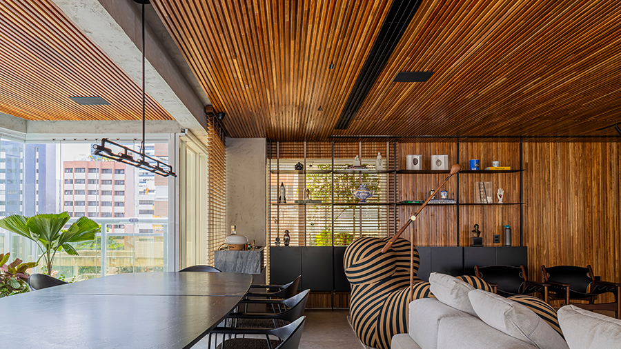 Ambiente com revestimento de teto e parede em brise eucalipto citri, com uma mesa rodeada de cadeiras. Um sofá com uma poltrona e ao fundo um armário aberto com objetos decorativos