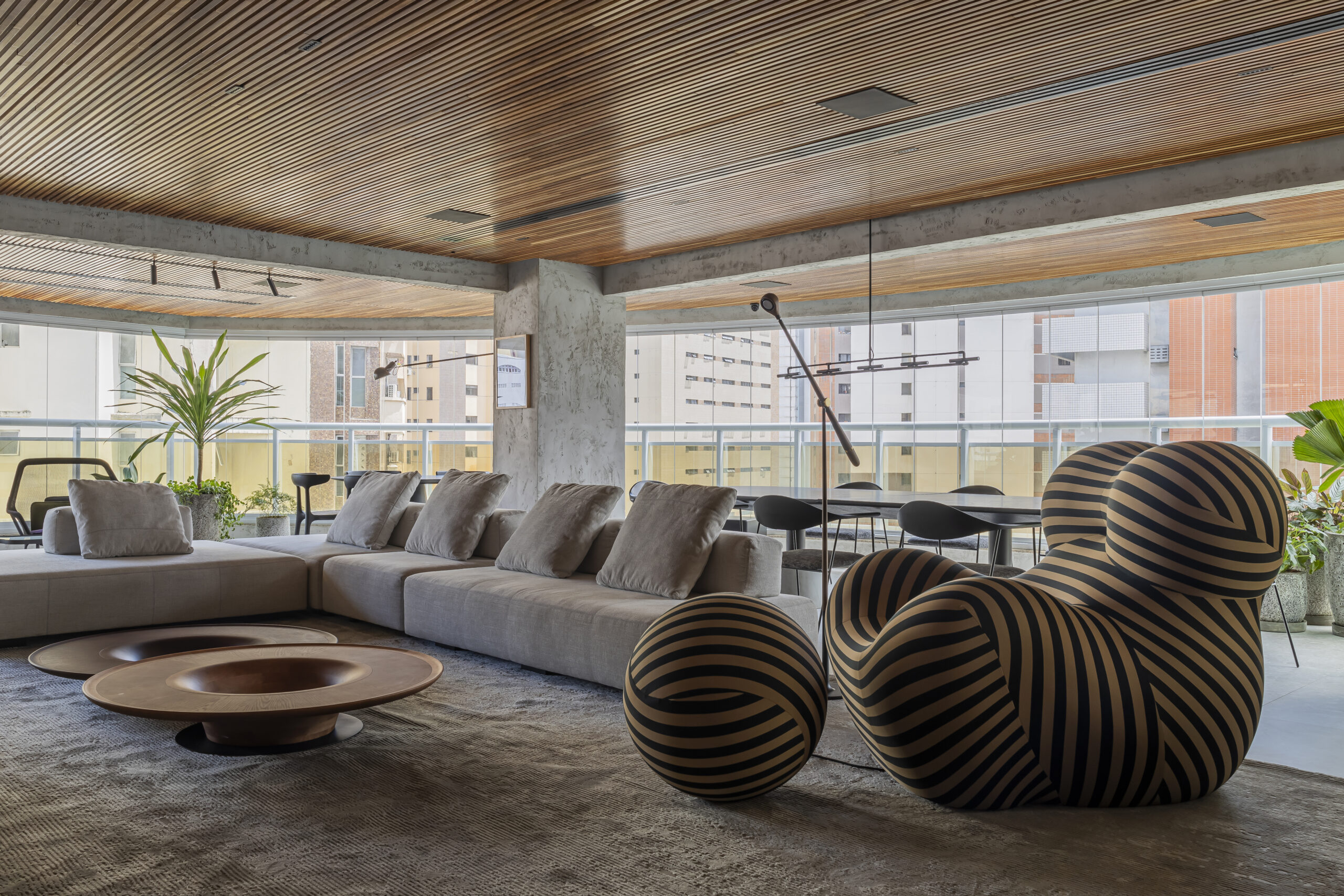 Imagem de uma sala com um sofá em L com almofadas, uma poltrona ao lado com um apoio para os pés e duas mesas de centro. O espaço ao redor possui uma sacada, e o teto é revestido com madeira brise eucalipto.