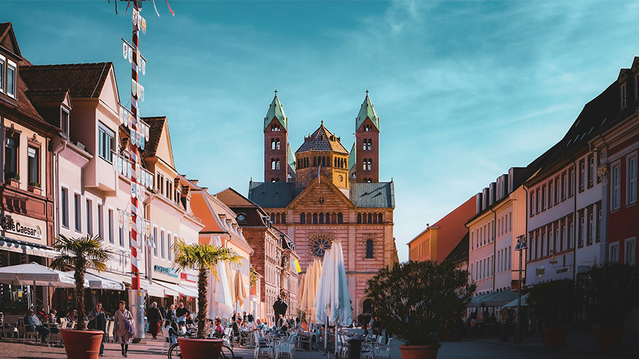 Imagem de uma cidade com um calçadão com árvores, mesas e pessoas. Nas laterais, prédios e ao fundo, uma igreja com arquitetura medieval românica.