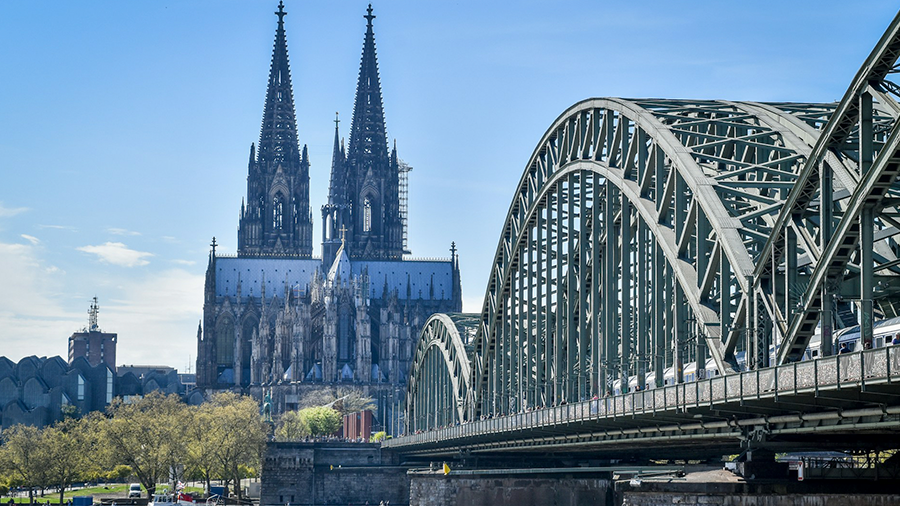 Imagem de um rio com um barco e acima uma grande ponte com estrutura em arcos. Ao fundo, diversas árvores e uma igreja com arquitetura gótica.