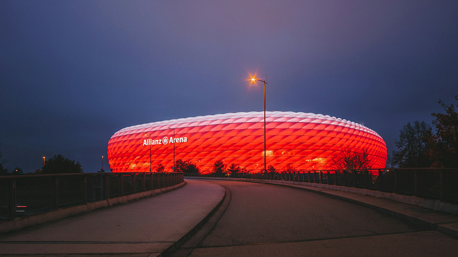 Imagem de uma rua com o estádio do Bayern de Munique ao fundo, com as luzes acesas na cor vermelha e o topo branco.