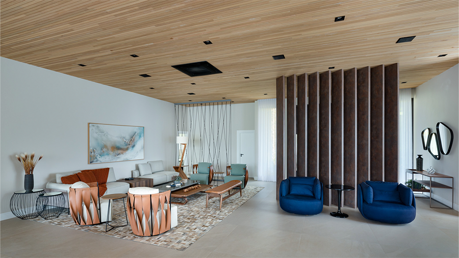  Imagem de uma sala com teto de madeira Lambri Tauari, poltronas, sofá, mesas de centro e laterais com objetos decorativos, um quadro na parede e uma das paredes revestida com madeira.