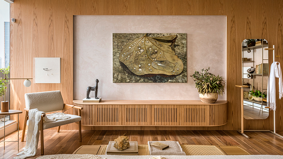 Imagem de uma sala com um rack, objetos decorativos em cima e um quadro pendurado na parede. De um lado, um espelho e do outro, uma poltrona com uma mesa com luminária, uma mesa de centro e um sofá, com o piso pronto de madeira de eucalipto citri.