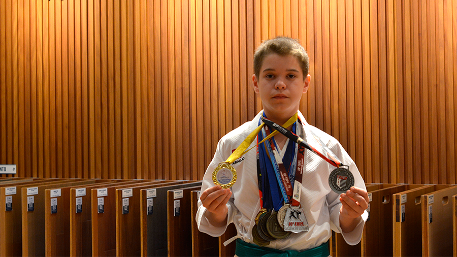  Imagem de Miguel Canavezi Stocco com roupa de karatê, medalhas no pescoço, segurando uma em cada mão e ao fundo um painel ripado de madeira com o logo e nome da Indusparquet.