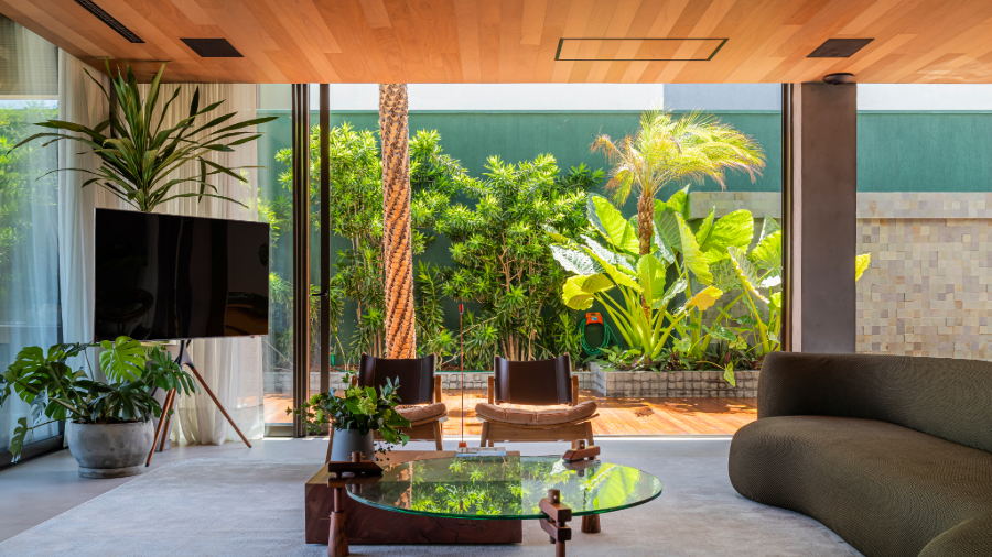 Imagem de uma sala com um sofá, duas mesas de centro sobre um tapete grande. À frente, uma TV com vasos ao lado e atrás. Na lateral, há uma grande janela que dá acesso a um espaço com plantas. O teto é de madeira MasterTeto Tauari Natural.