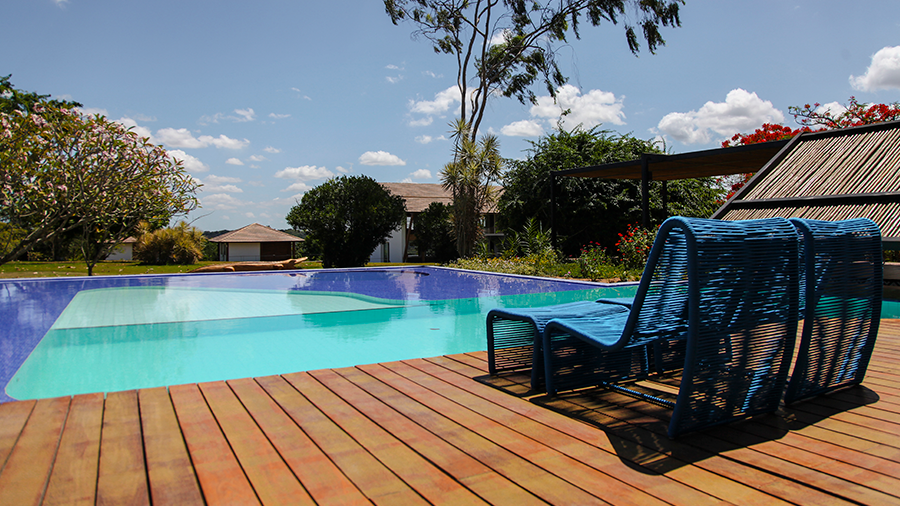 Imagem de uma área com piscina e um deck de madeira, com duas espreguiçadeiras azuis e várias árvores ao fundo.