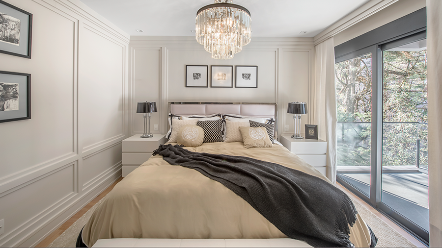 Quarto no estilo clássico, com moldura boiserie nas paredes, cortina branca na janela, cama no centro com duas mesas de cabeceiras, utilizando tons claros e cinza e um lustre de cristal acima da cama